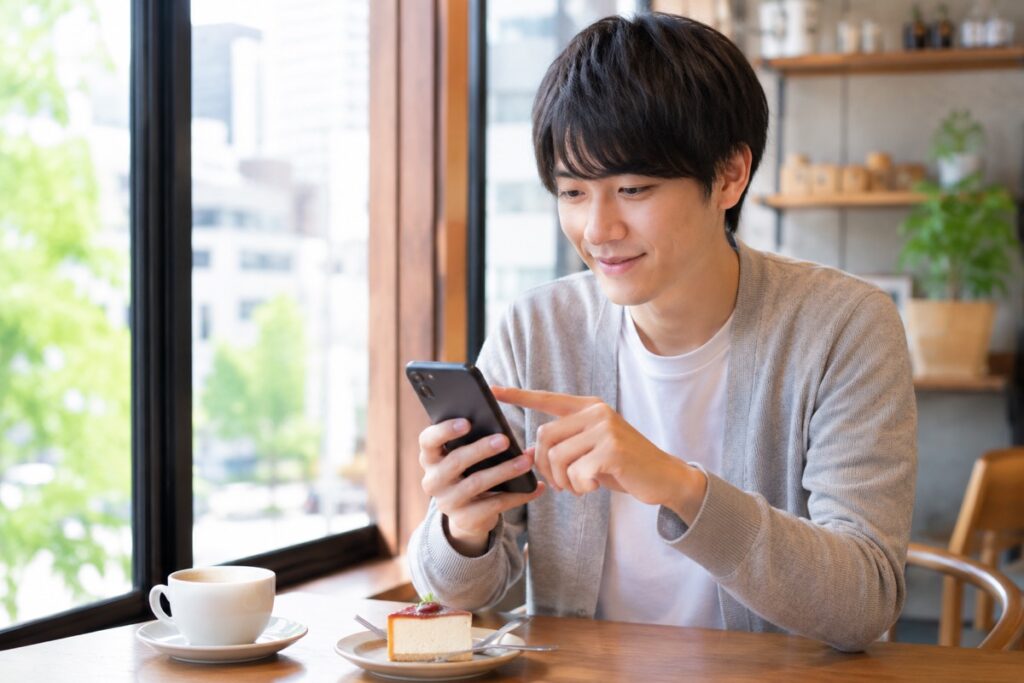 カフェの窓際でコーヒーとケーキを前にスマホを快適に操作する男性